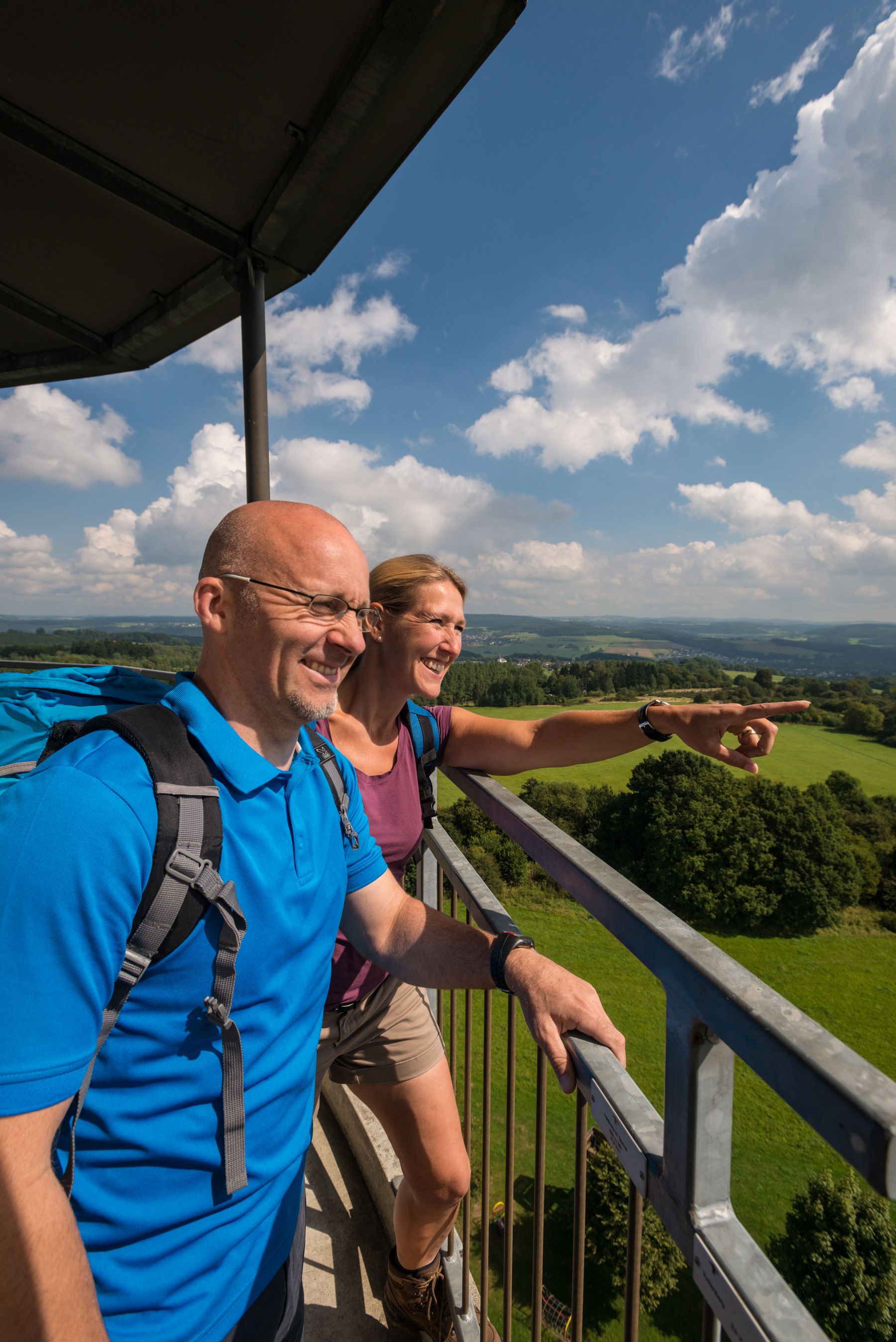 Ein Mann und eine Frau mit Rucksäcken stehen auf einer hohen Aussichtsplattform, lächeln und genießen die Aussicht auf grüne Felder und Wälder unter einem blauen Himmel mit Wolken. Die Frau deutet in die Ferne.
